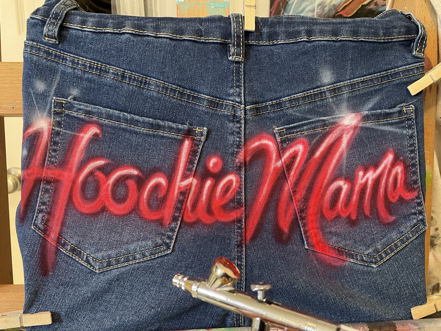 Red script lettering airbrushed across the back pockets of a denim piece, with the airbrush gun resting beside it, by Tina Cozart