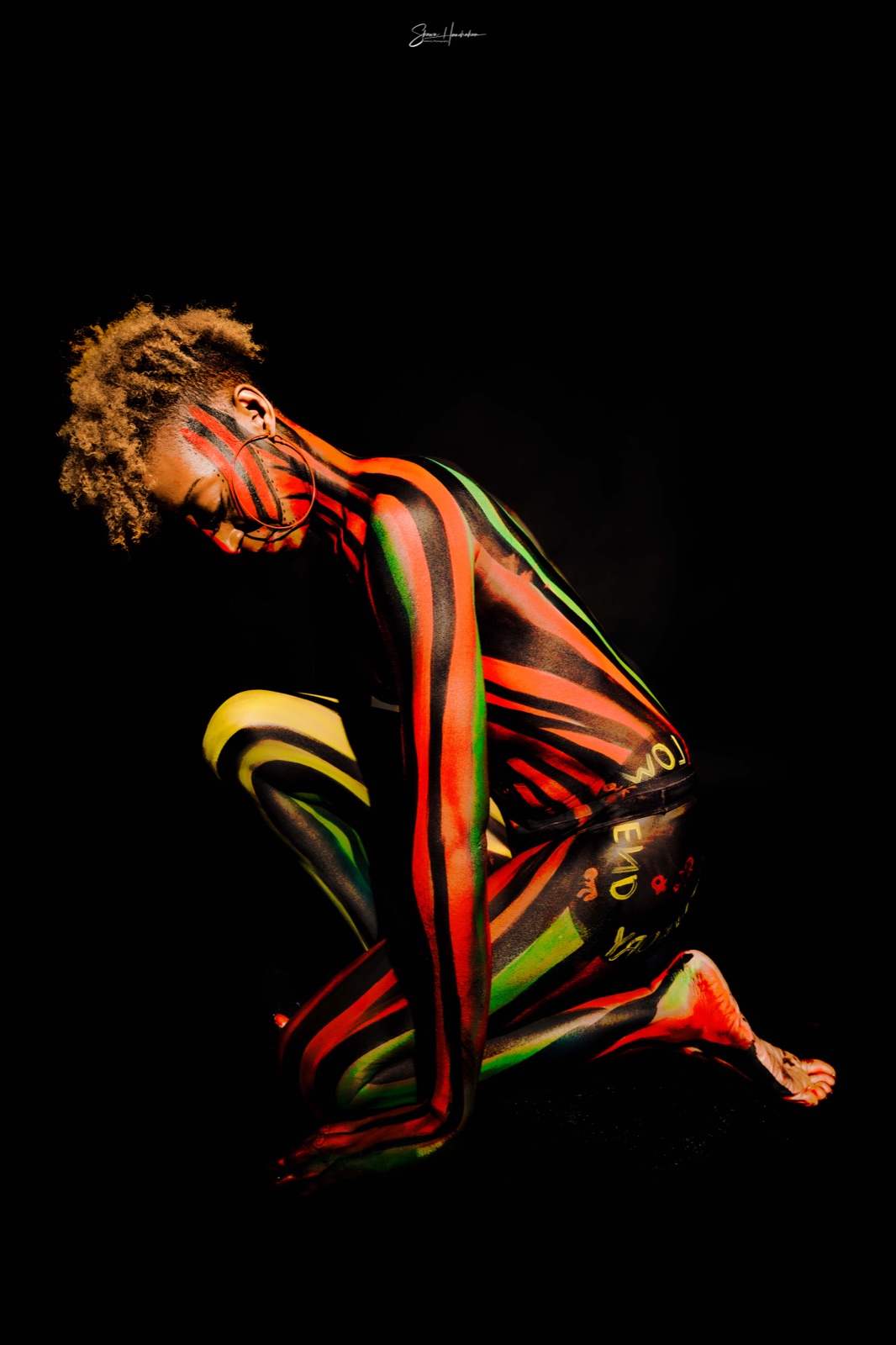 A kneeling figure in flame-colour UV body paint glowing on black