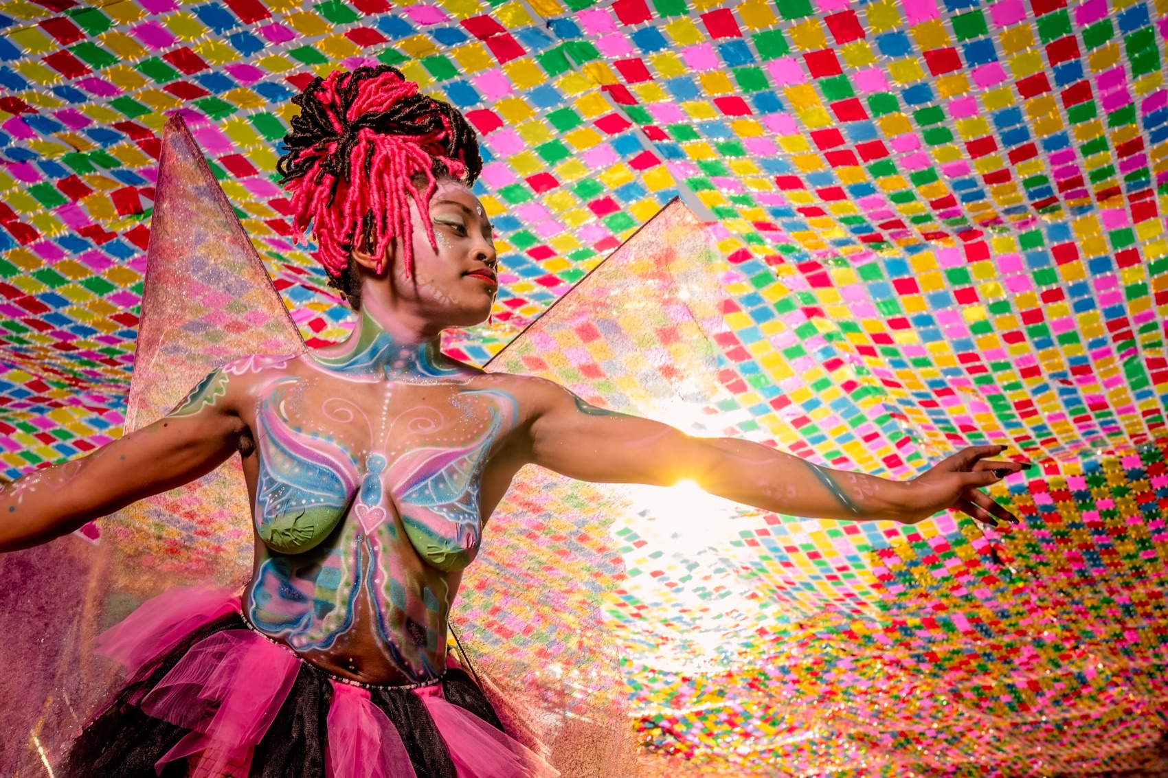 A model in vivid rainbow body and face paint, arms outstretched at a festival